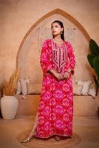 Woman in a pink patterned dress standing in a decorative room with an arch and plants.
