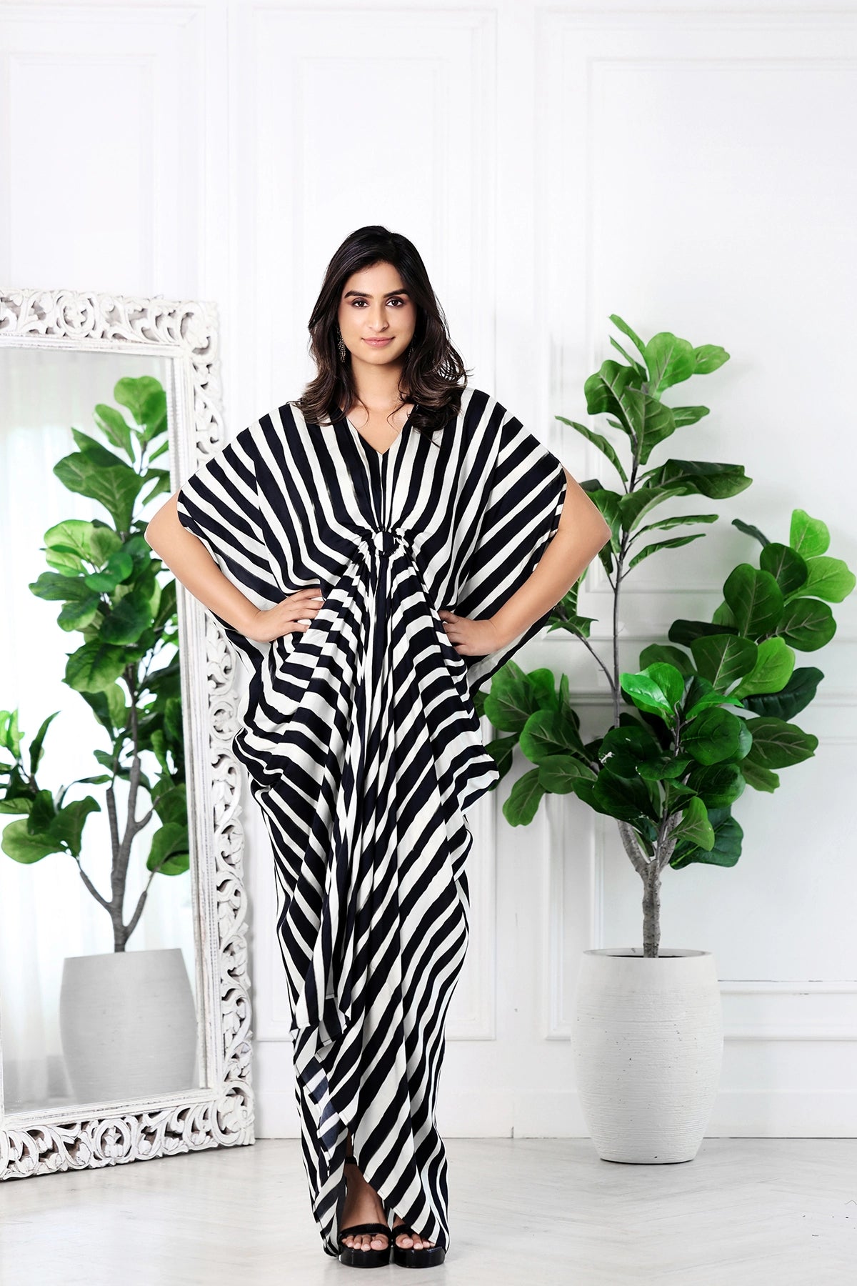 A woman modeling a black and white vertically striped kaftan dress in a bright, white-paneled room with potted fiddle-leaf fig plants and a large white-framed mirror.