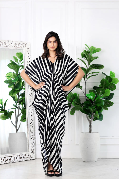 A woman modeling a black and white vertically striped kaftan dress in a bright, white-paneled room with potted fiddle-leaf fig plants and a large white-framed mirror.