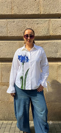 A woman wearing a white long-sleeve button-up shirt with a blue iris graphic on the front, paired with wide-leg blue jeans, standing outdoors against a beige stone wall.