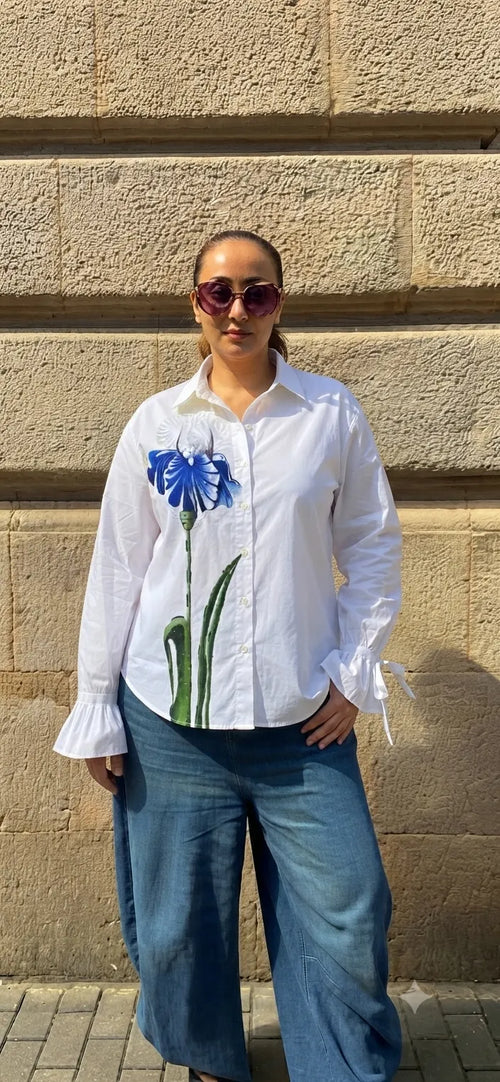 A woman wearing a white long-sleeve button-up shirt with a blue iris graphic on the front, paired with wide-leg blue jeans, standing outdoors against a beige stone wall.