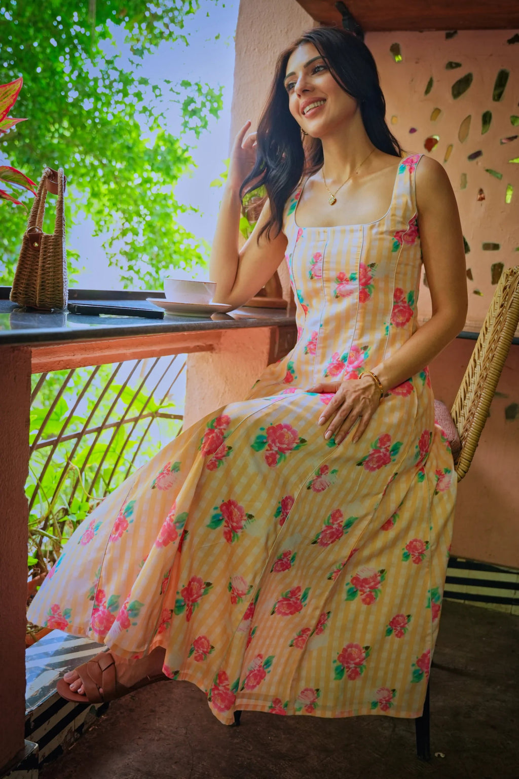 A woman wearing a sleeveless cream-colored maxi dress with a pink and green floral print, sitting on a chair on a balcony.