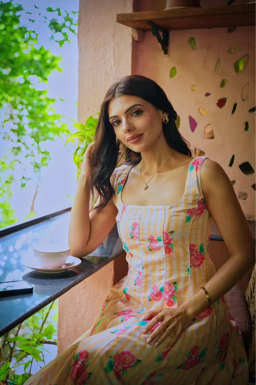 A woman wearing a sleeveless cream and yellow gingham dress with pink and green floral prints sits on a balcony next to a window, with a teacup on a saucer nearby.