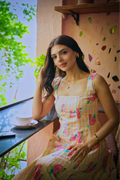 A woman wearing a sleeveless cream and yellow gingham dress with pink and green floral prints sits on a balcony next to a window, with a teacup on a saucer nearby.