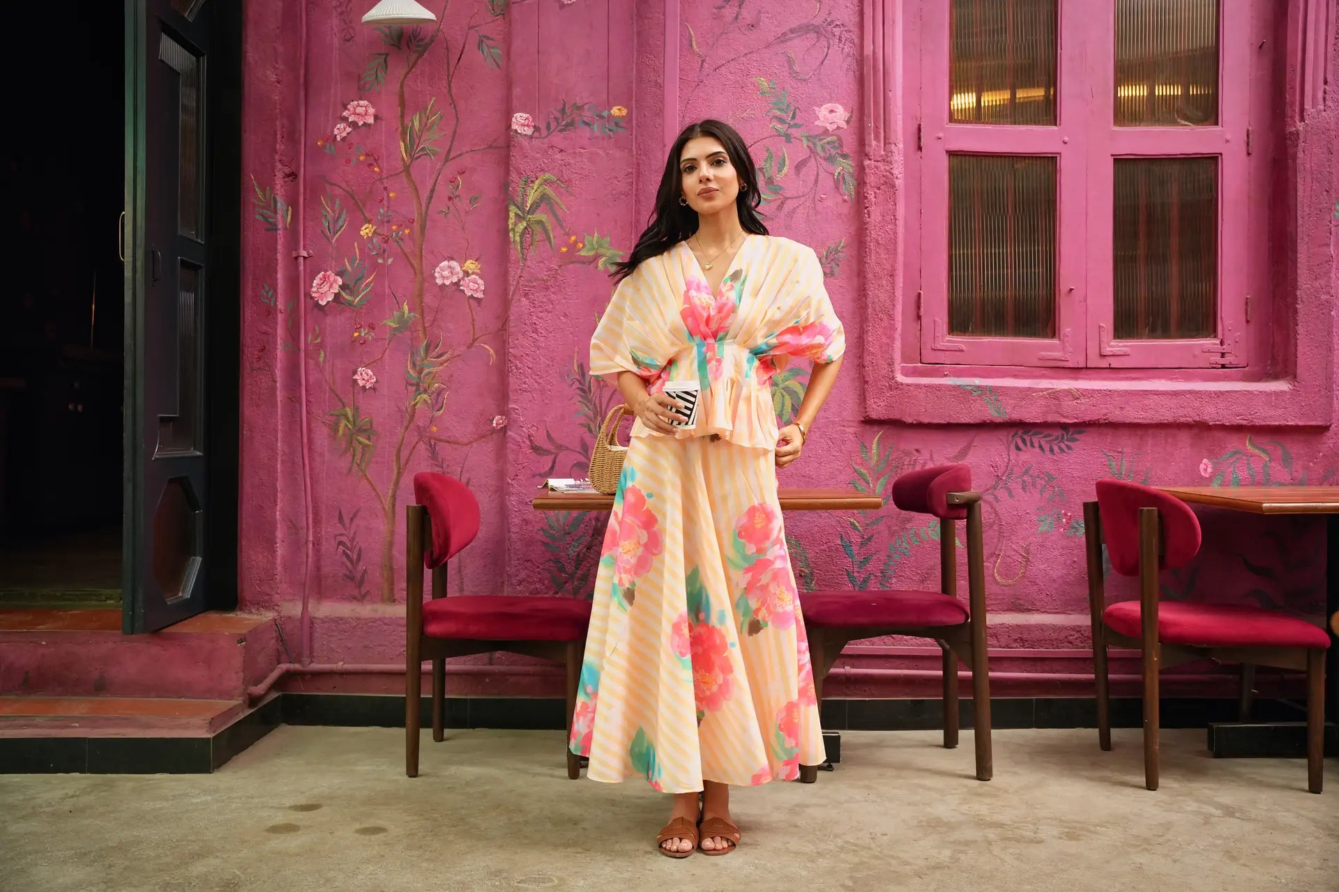 A woman stands in front of a vibrant pink floral wall, wearing a light beige kaftan dress with a colorful pink, green, and orange floral print. She is holding a small beige handbag.