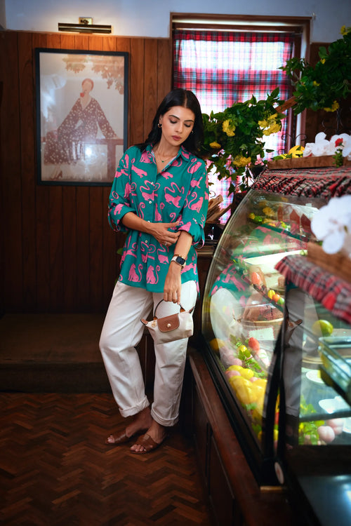 A woman stands in a bakery or cafe next to a glass display case, wearing a teal button-up shirt with a pink tropical print and white pants.