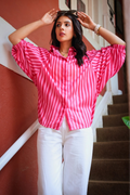 A woman wearing a vibrant pink and white vertically striped short-sleeve button-up shirt and white trousers, posing on a staircase with a terracotta-colored wall in the background.