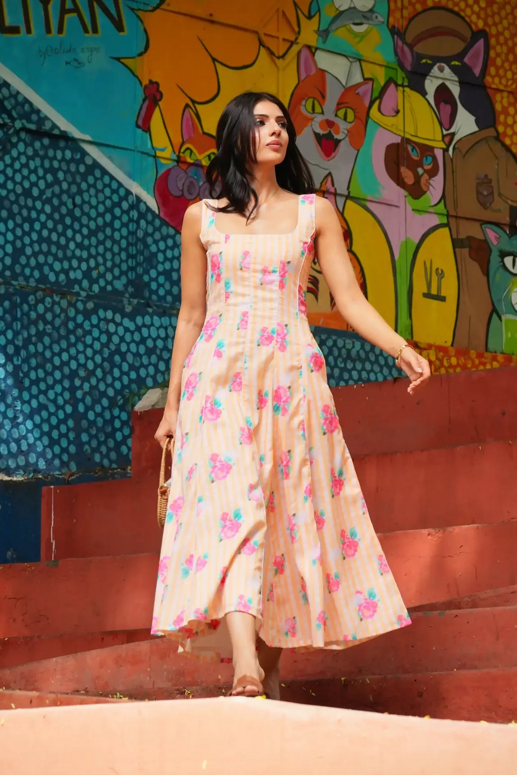 A woman wearing a sleeveless, light pink maxi dress with a colorful floral print walks down red concrete stairs in front of a vibrant, graffiti-style mural featuring cartoon animals.