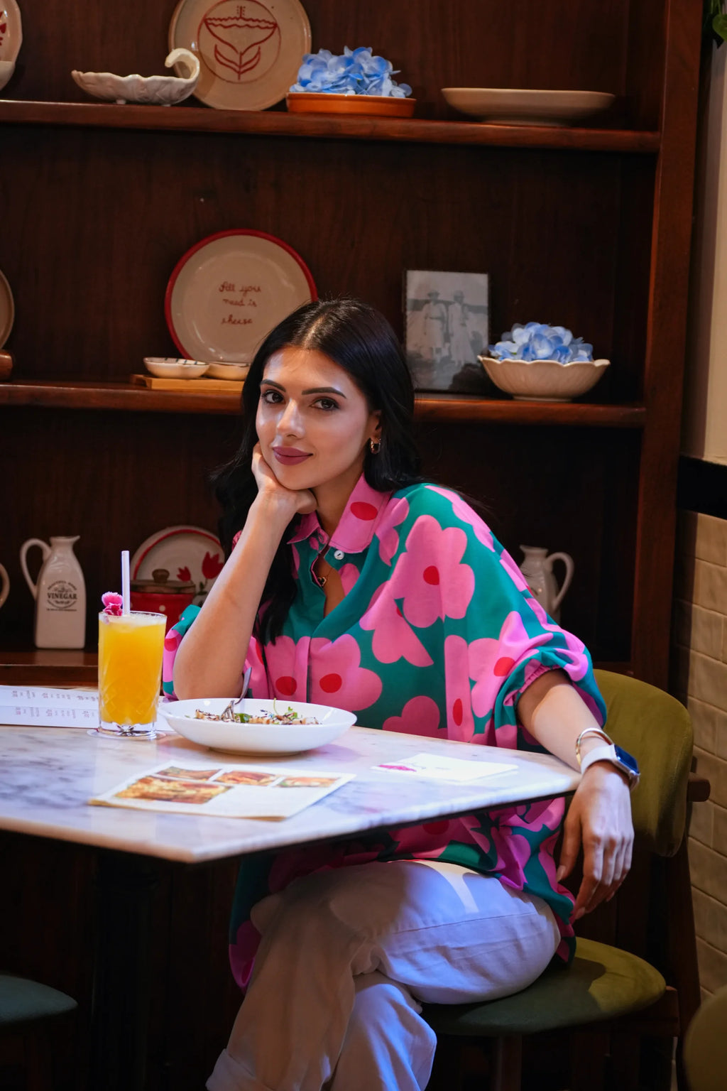 A woman sitting at a table in a cafe, wearing a vibrant green button-up shirt with large pink floral patterns and light beige trousers. A glass of orange juice and a plate of food are on the table in front of her.