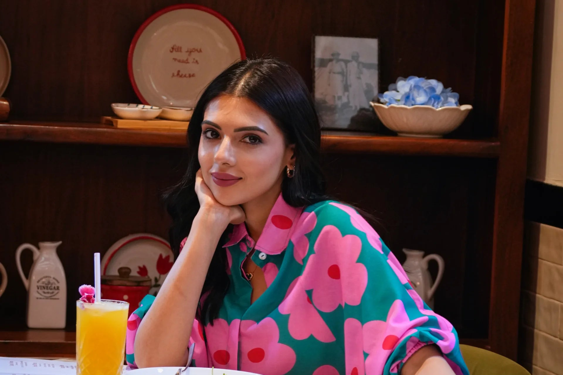 A woman with dark hair sitting at a table in a restaurant or cafe, wearing a vibrant green and pink floral print shirt. A glass of orange juice is in the foreground, and wooden shelves with decorative plates and bowls are in the background.
