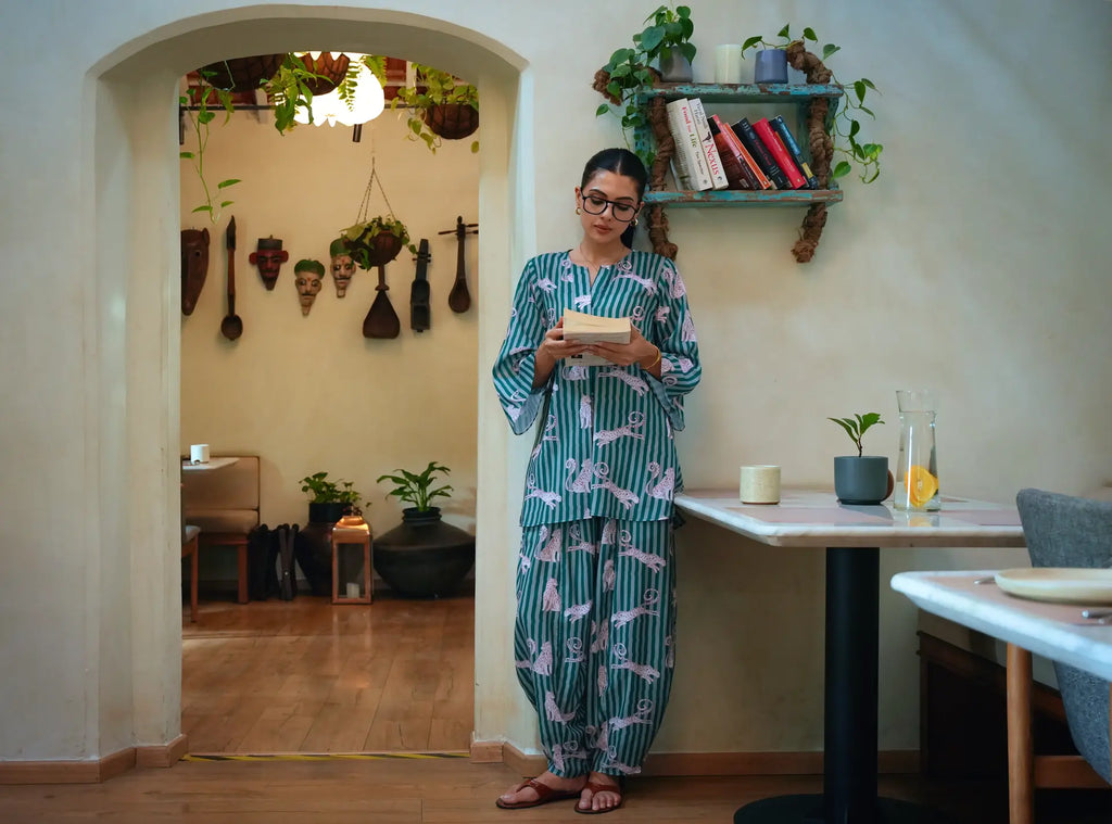 A woman wearing a matching green and white patterned pajama set stands in a cozy, warmly lit home interior, holding a small book or notebook. The room features an arched doorway, a wooden floor, a small table, and wall shelves with books and plants.