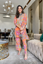 A woman standing in a living room wearing an orange traditional outfit with floral patterns and mirror work on the neckline.