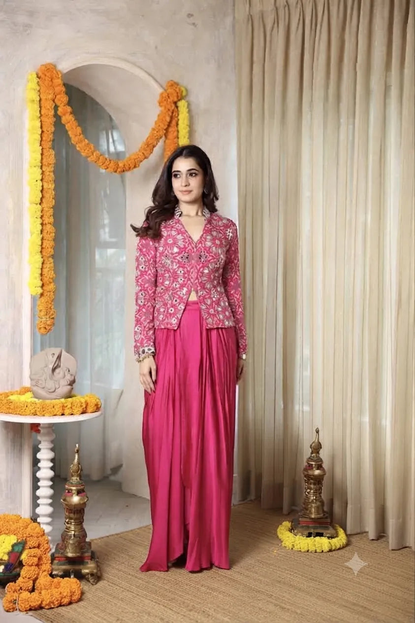 A woman modeling a traditional Indian outfit consisting of a pink floral embroidered jacket and a matching solid magenta skirt, standing in a decorated indoor setting with marigold garlands and beige curtains.
