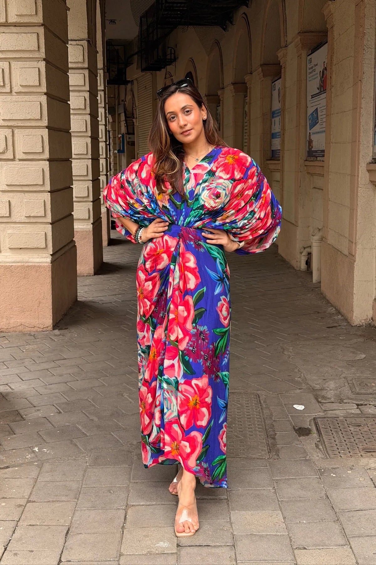 A woman wearing a vibrant, multicolored floral maxi dress with dramatic balloon sleeves, standing in a stone archway corridor.