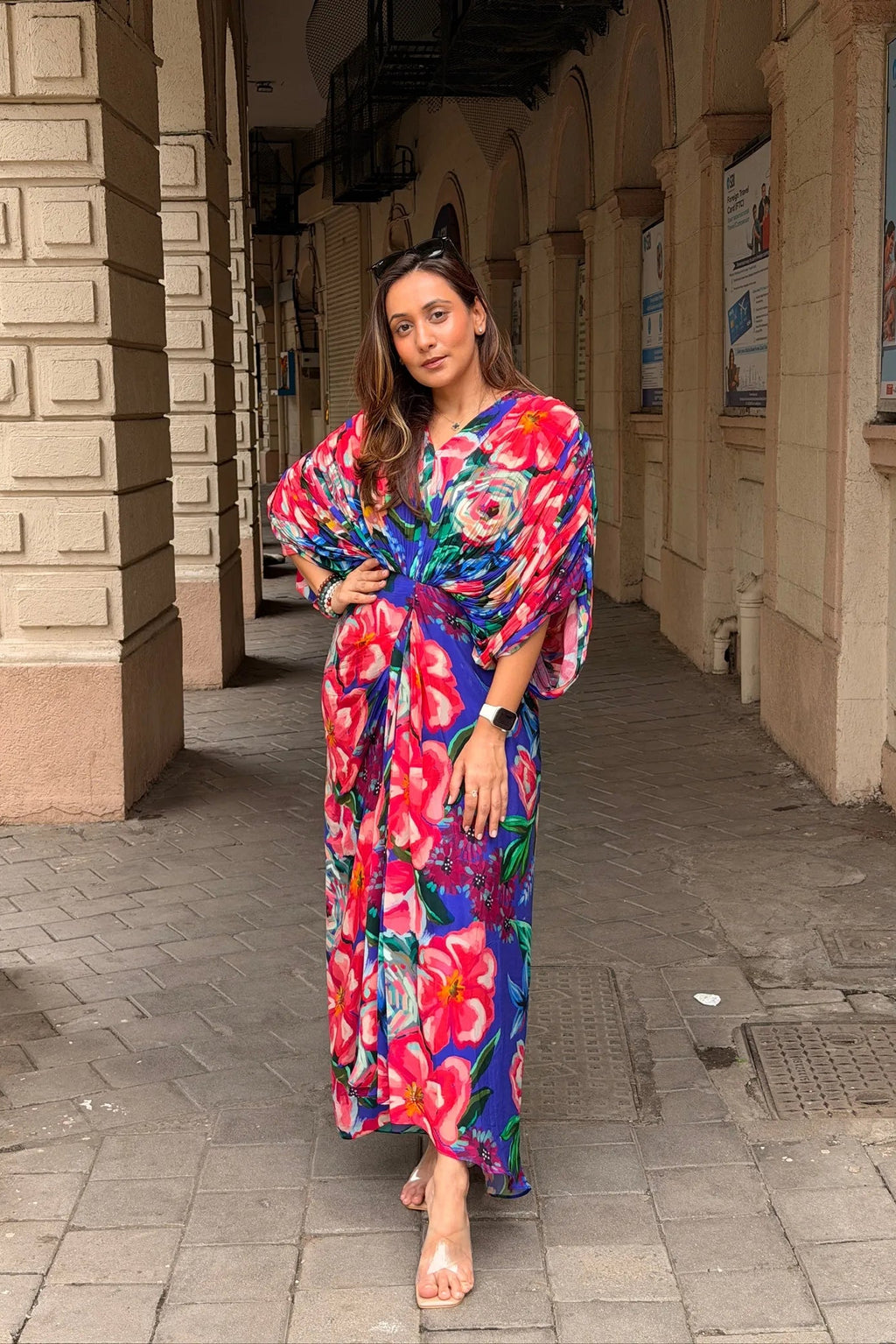 A woman stands in a covered outdoor walkway wearing a vibrant floral maxi dress with wide sleeves and a tie waist. The dress features a bold pattern of pink, blue, red, and green flowers on a purple base.