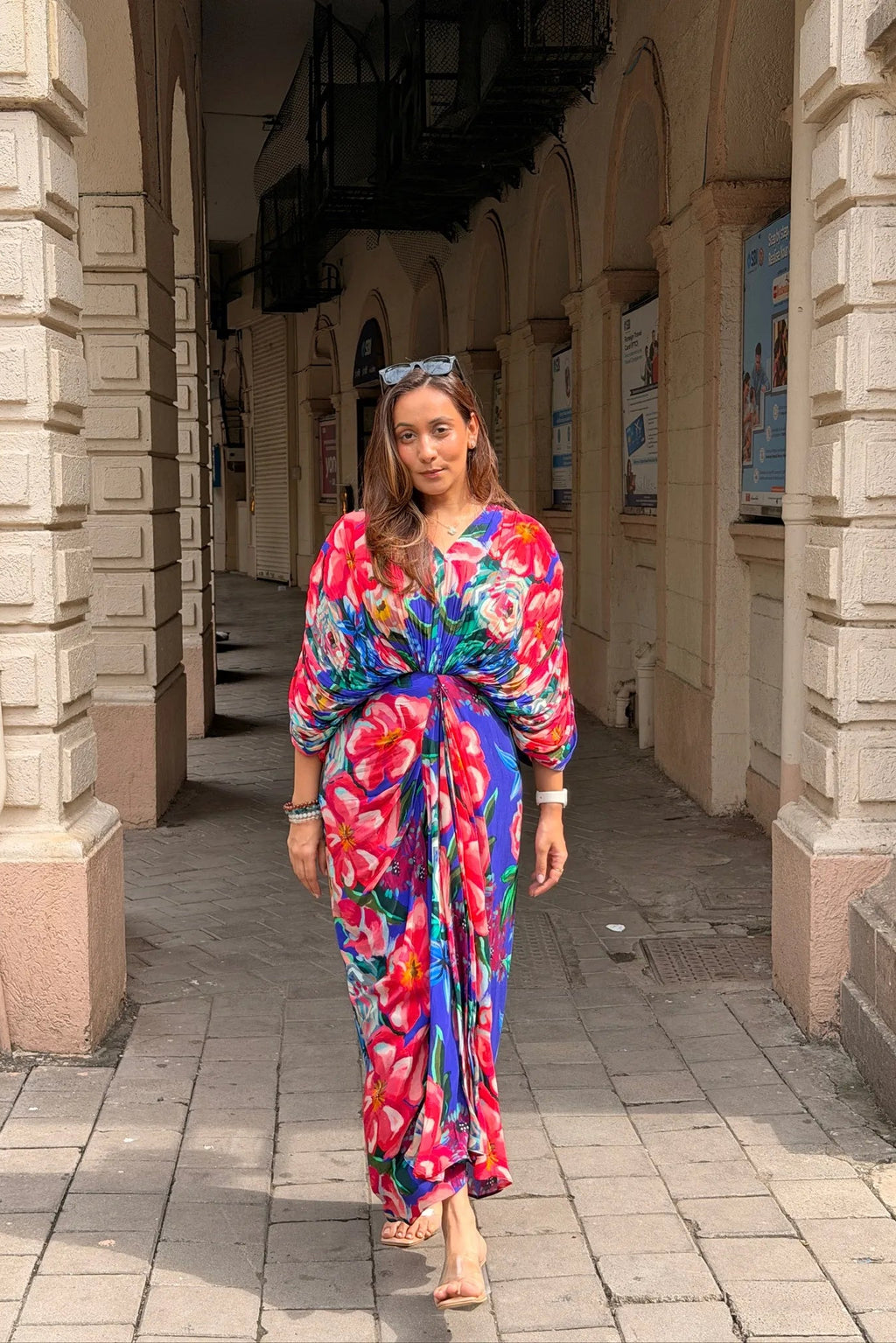 A woman walking through a stone arcade wearing a vibrant, multicolored floral print maxi dress with three-quarter sleeves and a self-tie waist.