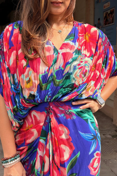 A woman wearing a vibrant, multicolored floral print kaftan dress with a wrap-style V-neck and gathered waist. The dress features a bold pattern of pink, red, blue, green, and white flowers.