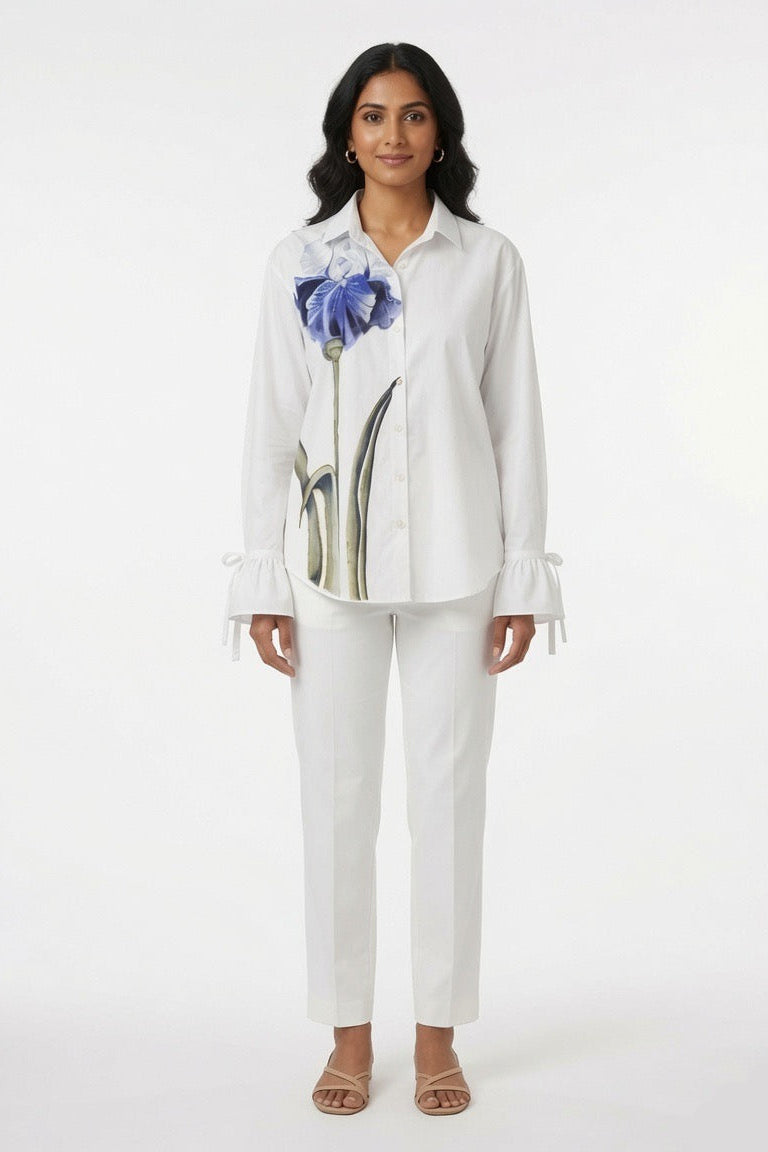 A woman modeling a white long-sleeve button-up shirt with a large blue and green floral graphic on the front, paired with white pants, against a plain white studio background.