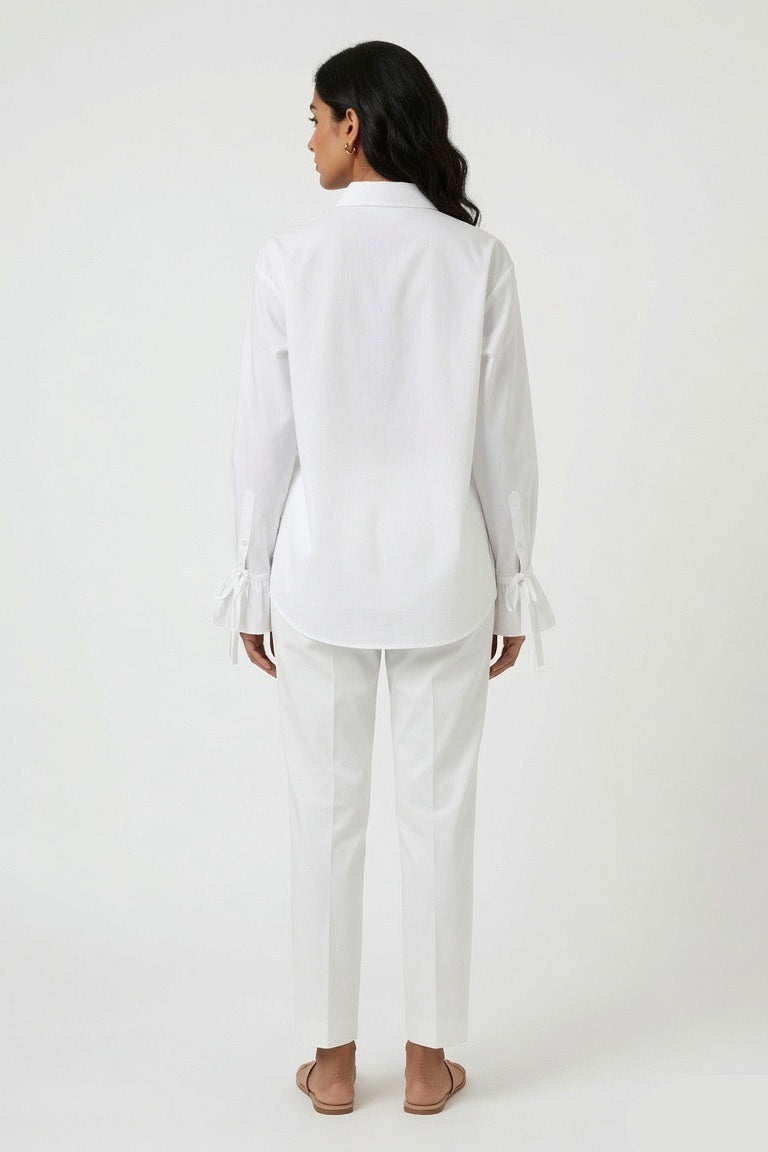 A woman shown from the back wearing a white long-sleeve button-up shirt with ruffled cuffs and matching white trousers, standing against a plain light gray studio background.