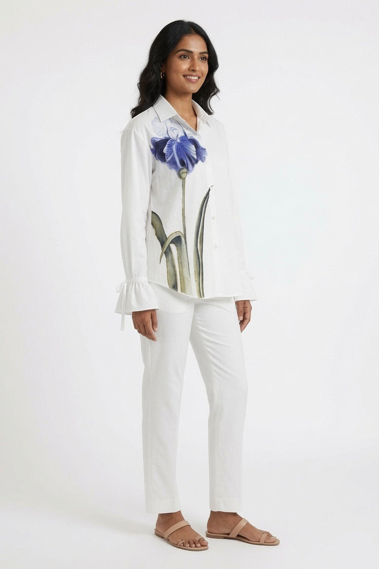A woman modeling a white long-sleeve button-up shirt with a large blue iris graphic on the front, paired with white pants and beige flat sandals, against a plain white studio background.