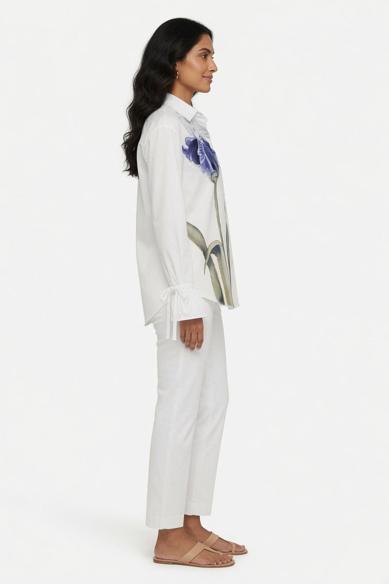 A woman shown in profile wearing a white long-sleeve collared shirt with a blue and gray abstract graphic print on the chest, paired with white cropped pants and tan flat sandals, against a plain white studio background.