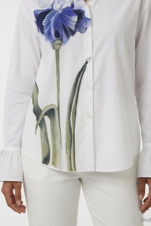 A white long-sleeve button-up shirt with a large blue and green iris flower graphic on the front, shown on a model wearing white pants against a light gray background.