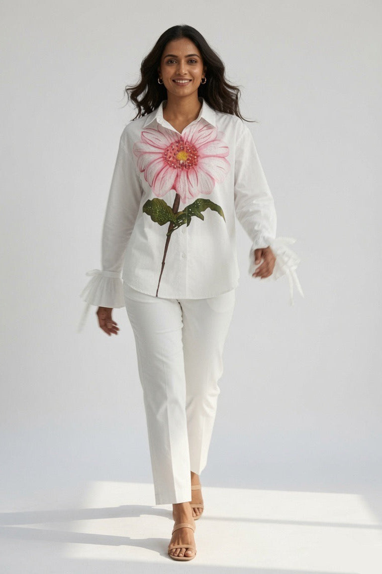 A woman modeling a white long-sleeve button-up shirt with a large pink and green flower graphic on the front, paired with white pants and tan heeled sandals, against a plain white studio background.
