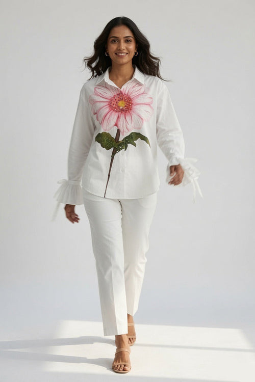 A woman modeling a white long-sleeve button-up shirt with a large pink and green flower graphic on the front, paired with white pants and tan heeled sandals, against a plain white studio background.