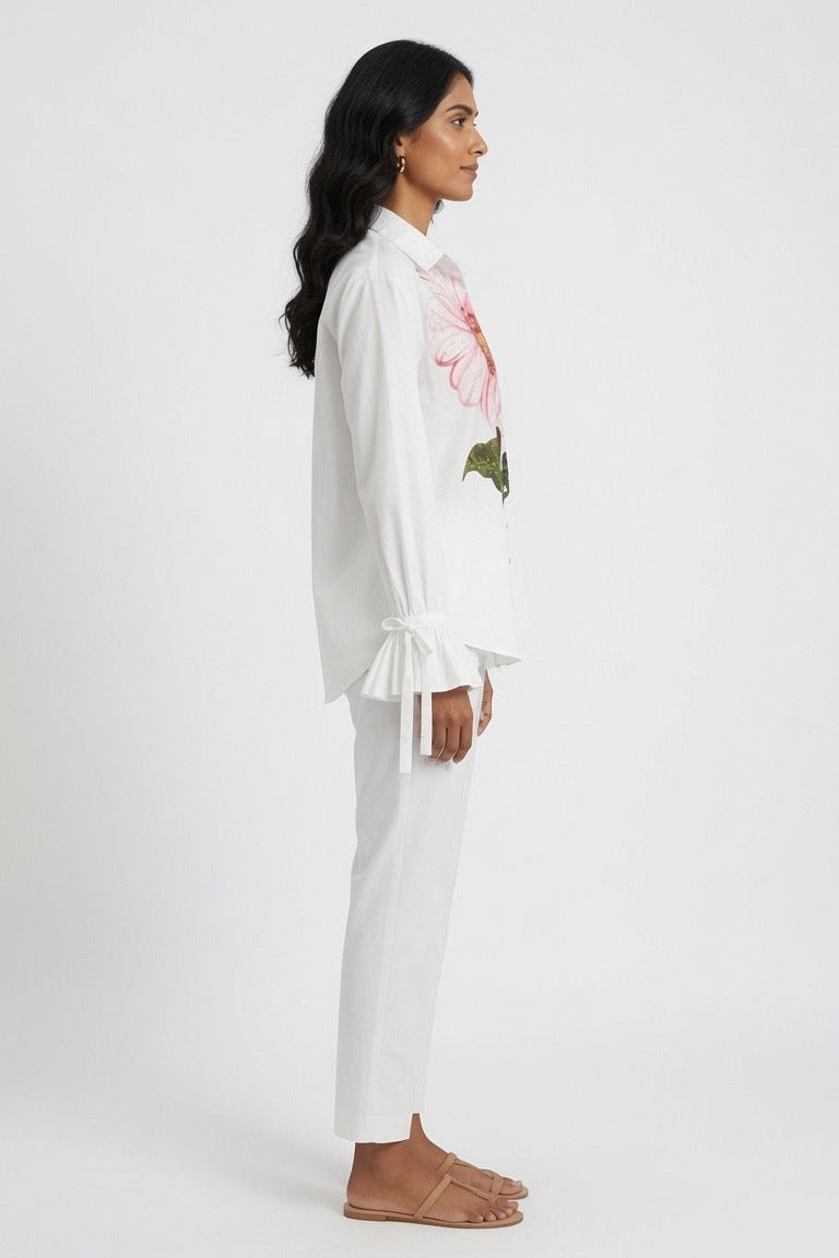 A woman shown in side profile wearing a white long-sleeve blouse with a pink floral and green leaf print on the chest, paired with white pants and tan flat sandals, against a plain light gray studio background.