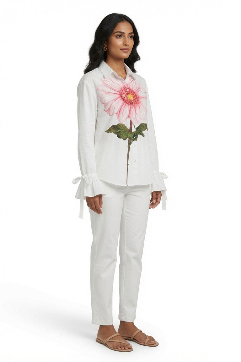 A woman modeling a white two-piece linen set featuring a long-sleeve button-up shirt with a large pink and yellow flower graphic on the chest, and matching white trousers, set against a plain white background.
