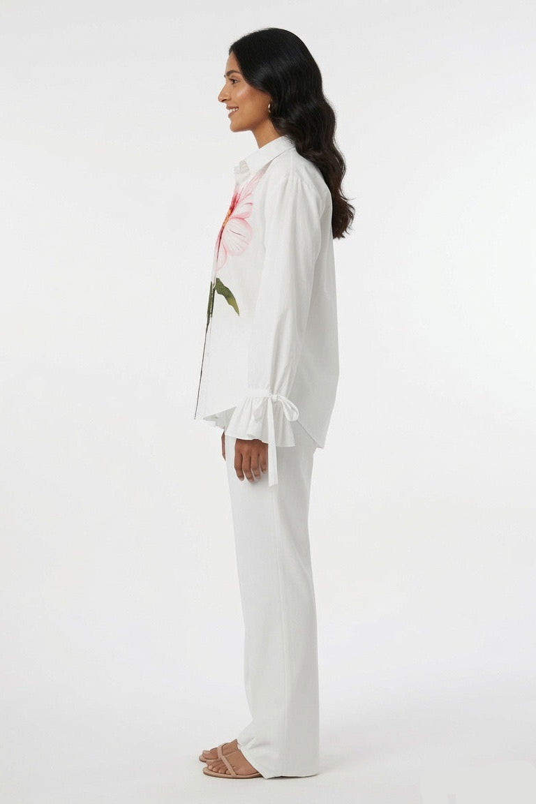 A woman shown in profile wearing a white long-sleeve blouse with pink floral embroidery and tie details at the cuffs, paired with white wide-leg pants, against a plain white studio background.