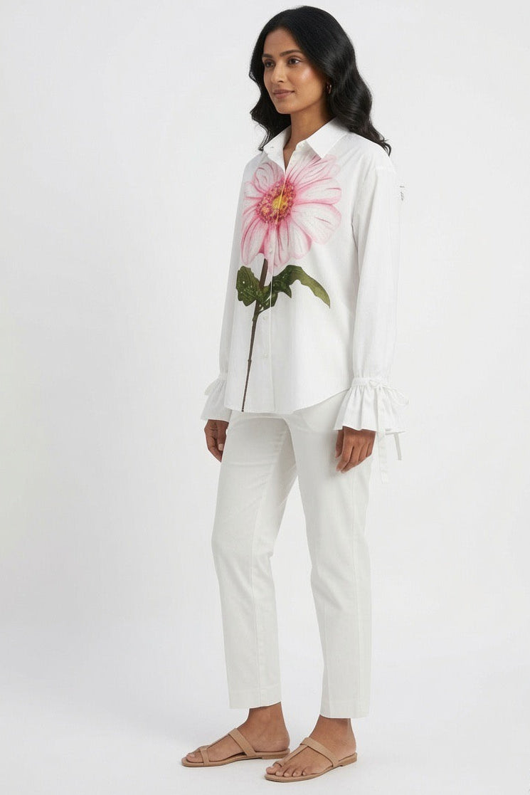 A woman modeling a white long-sleeve button-up shirt with a large pink and green flower graphic on the chest, paired with white pants and tan flat sandals, against a plain white studio background.