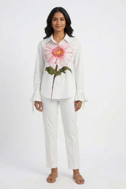 A woman modeling a white long-sleeve button-up shirt with a large pink and yellow flower graphic on the front, paired with white pants and tan sandals, against a plain white studio background.