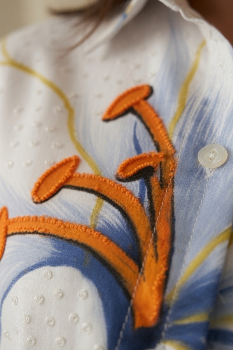 Close-up of a white button-up shirt with blue and yellow abstract brushstroke patterns and orange embroidered floral or branch-like applique.
