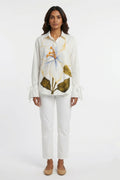 A model stands against a plain white studio background wearing a white long-sleeve button-up shirt with a large floral print in blue, gold, and olive green, paired with white cropped pants and beige flats.
