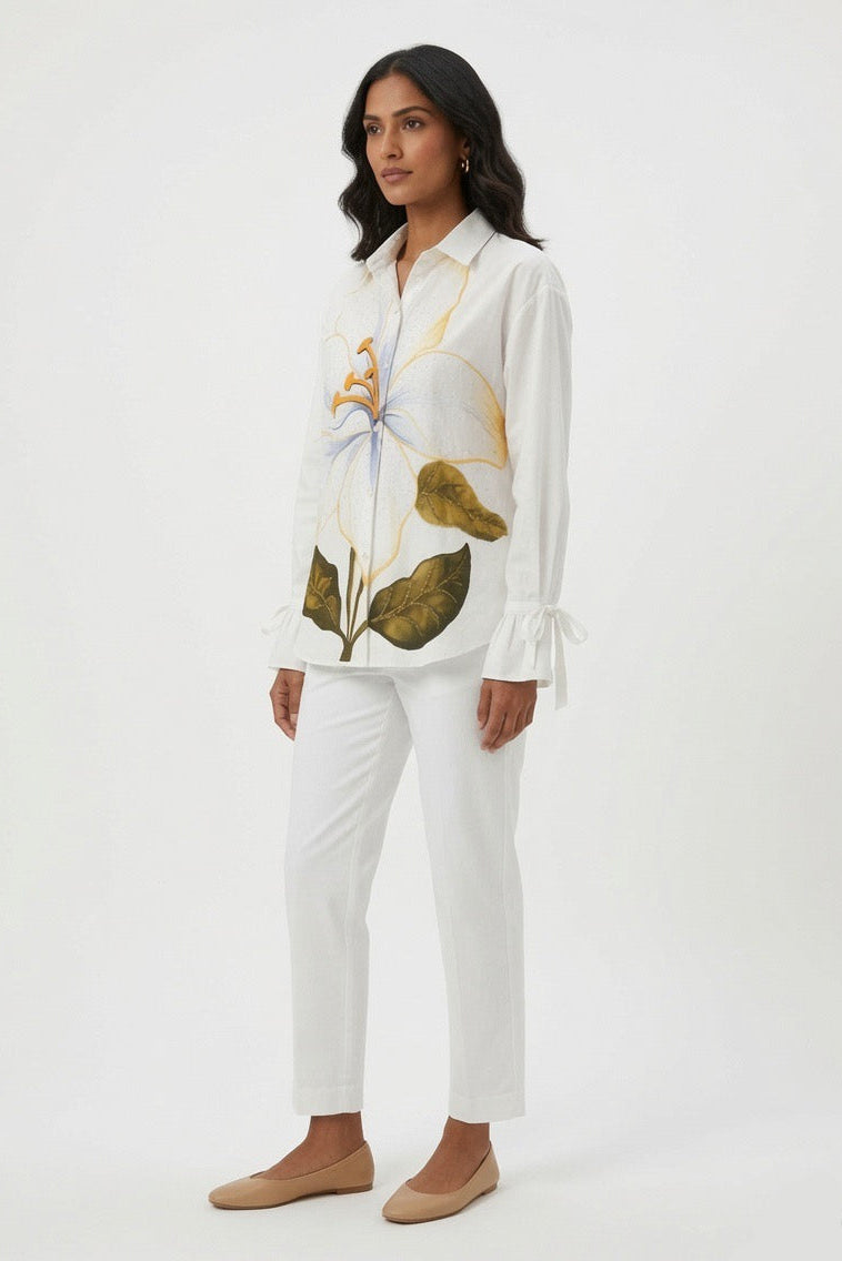 A woman modeling a white long-sleeve button-up shirt with a large floral print in yellow, green, and blue, paired with white cropped pants and beige flats, against a plain white studio background.