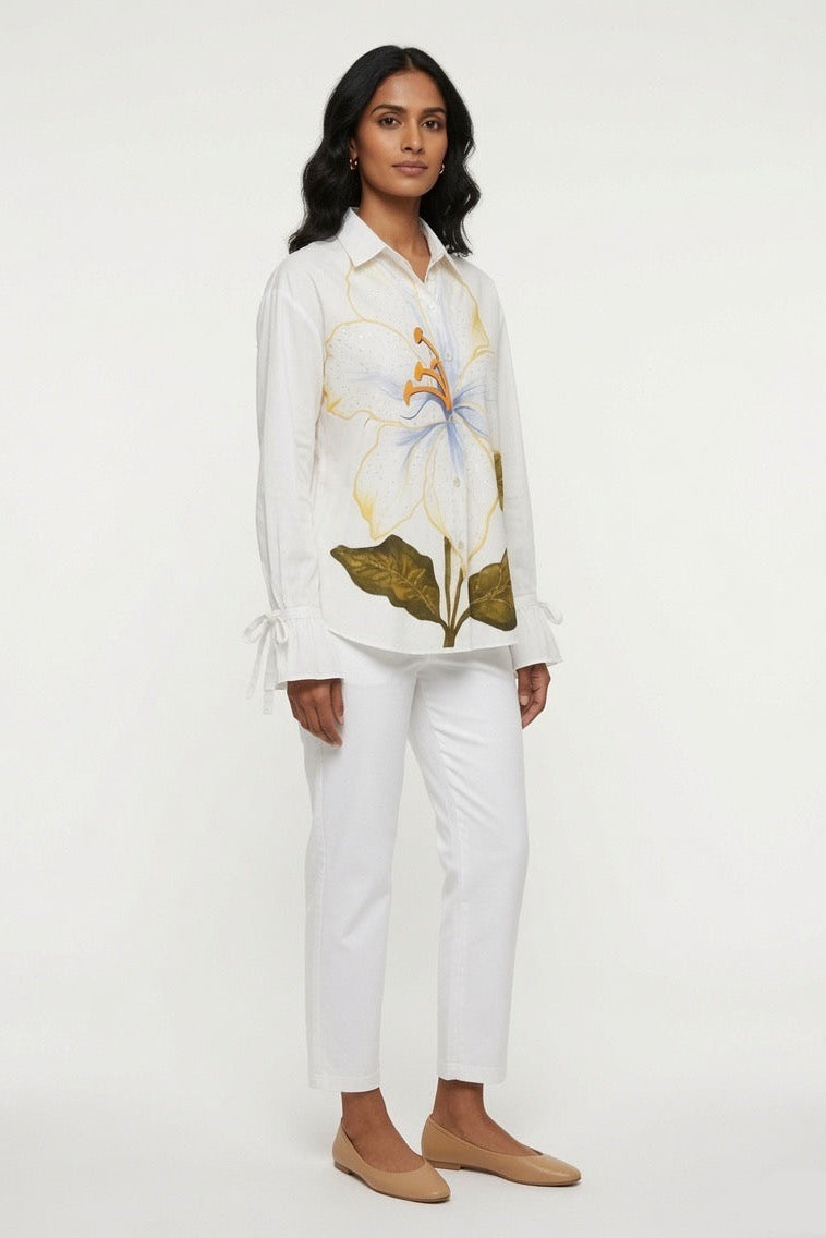 A woman modeling a white long-sleeve button-up shirt with a large blue and gold floral print on the front, paired with white cropped pants and tan flats, against a plain white studio background.