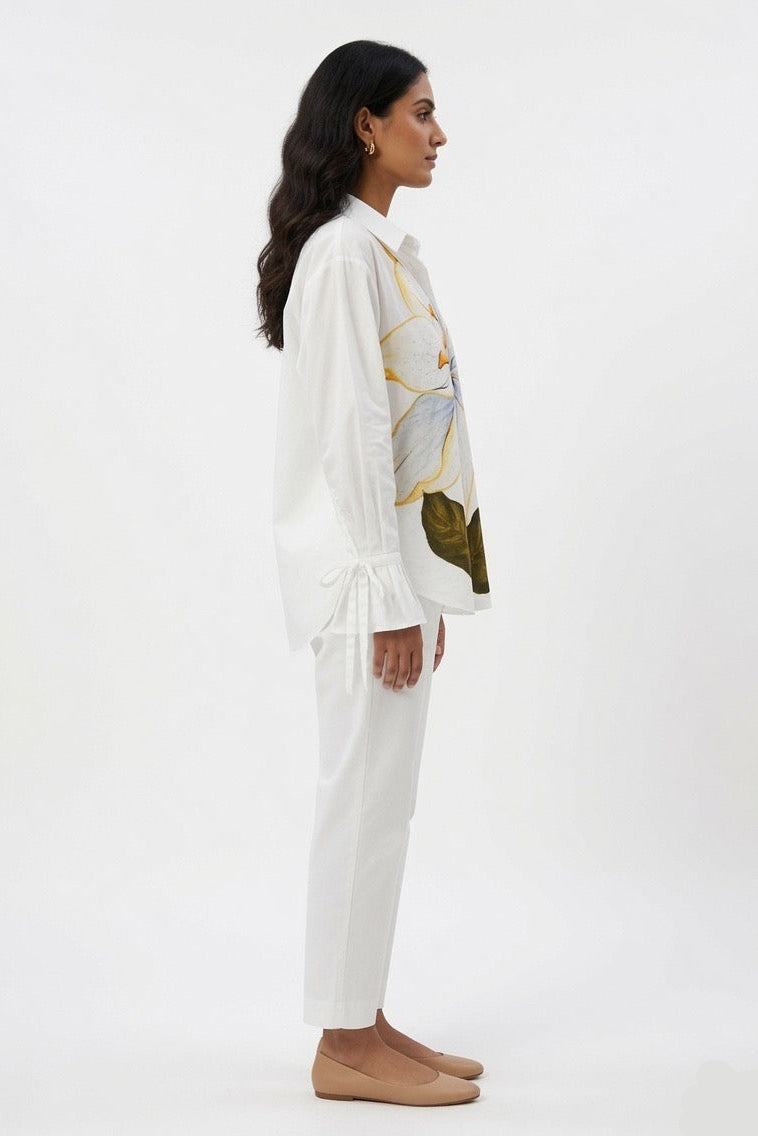 A woman shown in profile wearing a white long-sleeve blouse with a gold, blue, and olive abstract print on the front, paired with white trousers and tan flats, against a plain white studio background.