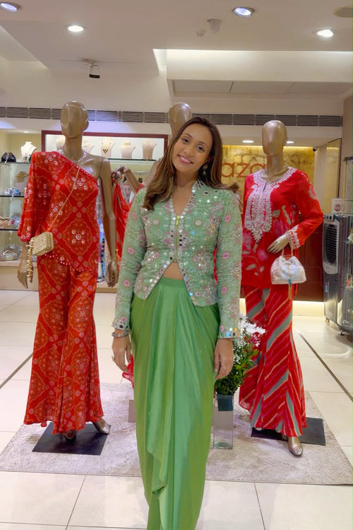 A woman wearing a light green embroidered kurta and matching green pants stands in a clothing store between two mannequins dressed in red traditional outfits.
