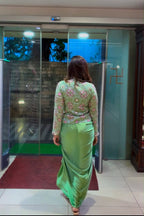 A woman viewed from behind wearing a light green traditional outfit with a patterned top and a flowing skirt, standing in front of glass doors at what appears to be a building entrance.