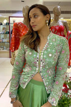 A woman wearing a light green embroidered jacket with pink and white floral patterns and a matching green skirt, standing in a clothing store with mannequins in the background.