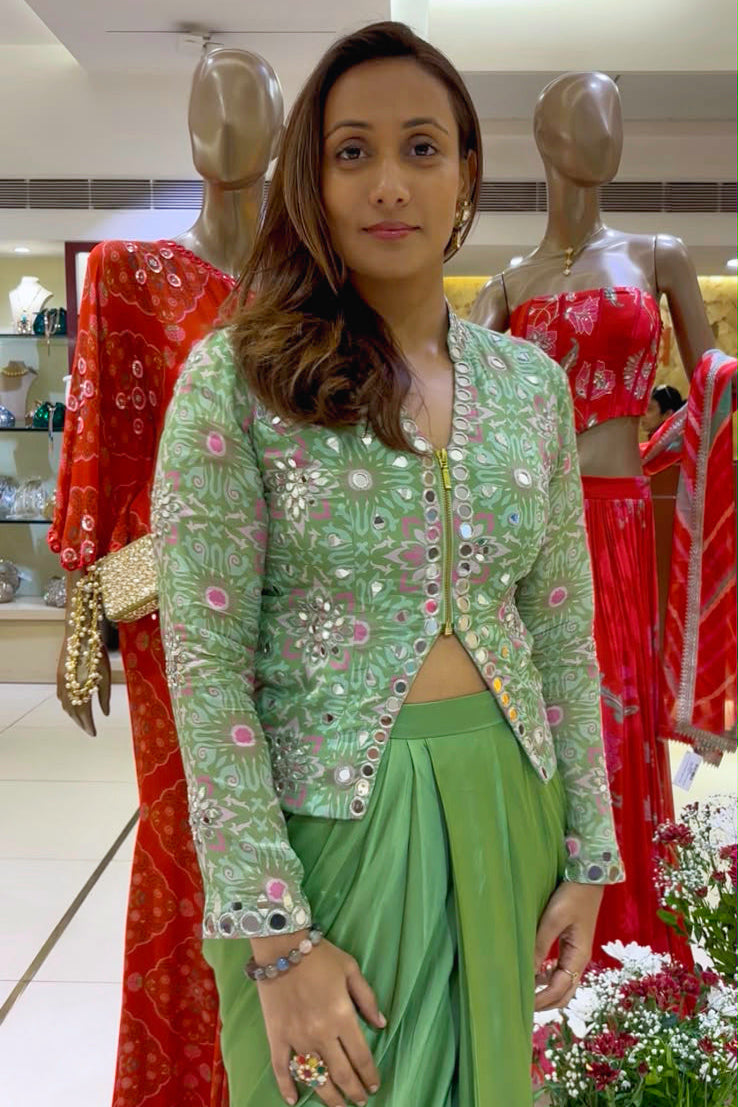 A woman modeling a light green embroidered jacket with a matching green skirt, standing in a clothing store with mannequins wearing red outfits in the background.