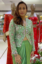 A woman modeling a light green embroidered jacket with a matching green skirt, standing in a clothing store with mannequins wearing red outfits in the background.