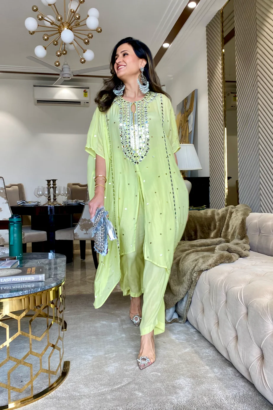 A person wearing a pastel green kaftan and pants set with intricate prints, standing in a stylishly furnished room.