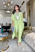 A person wearing a pastel green kaftan and pants set with intricate prints, standing in a stylishly furnished room.