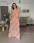 A woman modeling a sleeveless, floor-length halter dress with a pink, orange, and yellow floral print on a light beige background, standing in a living room.
