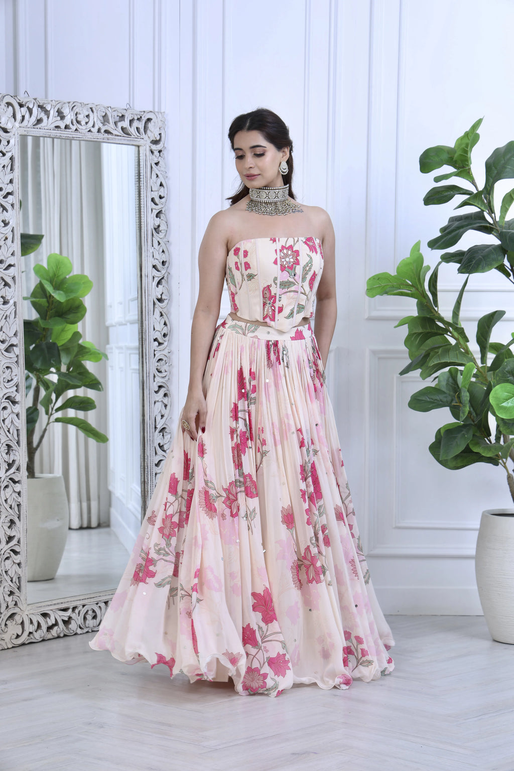 A woman models a strapless floral print gown with a fitted bodice and a full pleated skirt. The dress features a pink and red floral pattern on a light beige base.