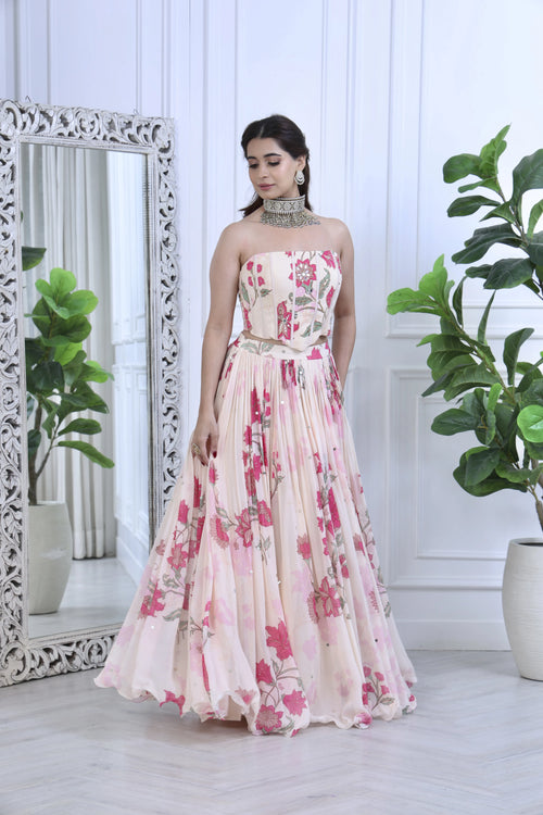 A woman models a strapless floral print gown with a fitted bodice and a full pleated skirt. The dress features a pink and red floral pattern on a light beige base.