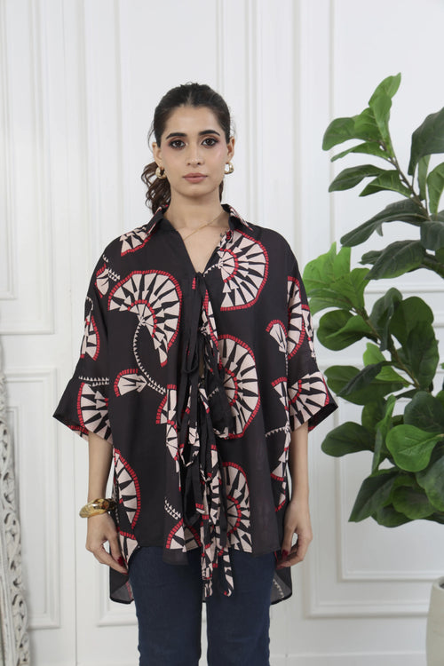 A woman modeling a black short-sleeve tie-front blouse with a beige and red circular paisley pattern, paired with dark blue jeans, standing in front of a white paneled wall with a green plant on the right.